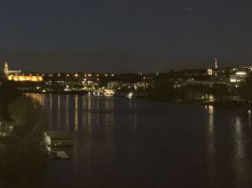 A serene cityscape at night with illuminated buildings reflecting on a calm river, dark sky above, and faint lights outlining the riverbanks and architecture. Peaceful atmosphere.