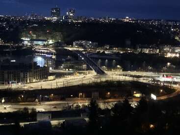 This nighttime cityscape features illuminated buildings, a bridge over a river, and roads with sparse traffic. The city lights create a vibrant urban atmosphere.