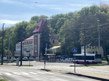 A historic building with a red roof sits beside a tree-lined street. Parked cars, a city bus, and a bus stop are visible in the foreground.