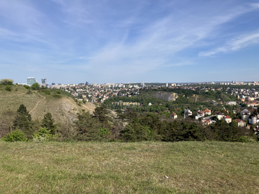 The image shows a scenic grassy hilltop overlooking a city with scattered houses, modern buildings, lush greenery, and a clear blue sky with wispy clouds.