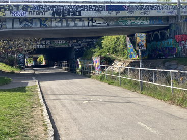 A paved path runs under two graffiti-covered bridges. Green grass and bushes line the sides. Sunlight and shadows create a vibrant, urban atmosphere with colorful street art.