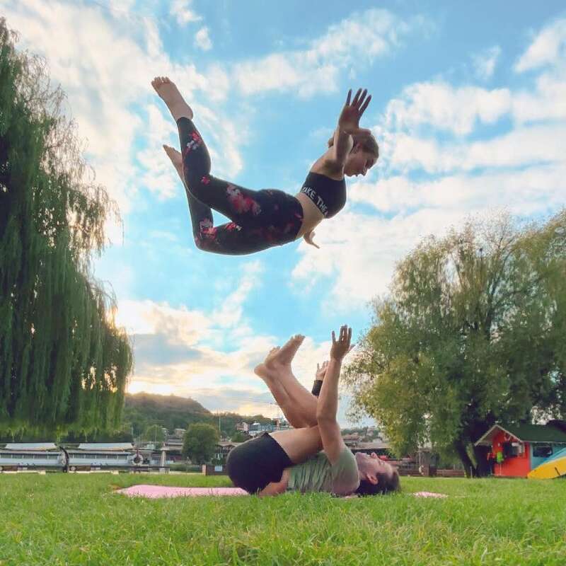 Two people practice acro yoga on the grass. One person lies on their back holding up the other, who is airborne against a bright, blue sky.