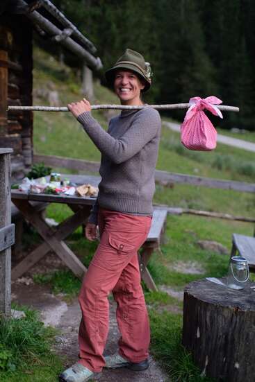 Une personne souriante, vêtue de vêtements de plein air et coiffée d'un chapeau vert, tient un bâton sur son épaule avec un paquet de tissu rouge noué, debout près d'une table de pique-nique rustique.