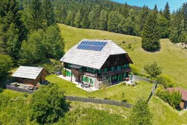 Une grande maison en bois avec un toit en panneaux solaires se trouve sur une colline herbeuse, entourée d'arbres, d'une petite cabane, de jardins et d'une clôture en bois rustique.