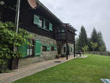 Une maison rustique en pierre et en bois avec des volets verts se trouve à côté d'une pelouse bien entretenue. Des arbres et des meubles d'extérieur complètent ce cadre paisible et champêtre sous un ciel couvert.