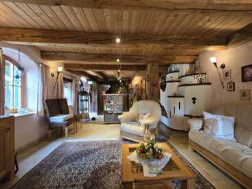 Ce salon confortable est doté de poutres en bois, d'un plafond rustique, de canapés beiges, d'un tapis à motifs, d'un poêle en faïence, d'un éclairage doux et de charmants détails décoratifs.