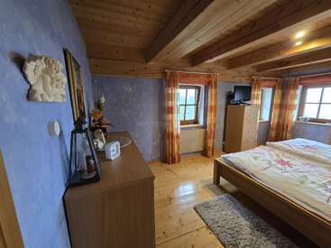Une chambre à coucher confortable avec des poutres en bois apparentes, des murs violets clairs et un plancher en bois. Elle comprend un lit, une commode, une télévision, des éléments décoratifs, des fenêtres et des rideaux à carreaux orange.