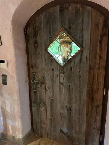 Une porte rustique en bois avec une fenêtre en verre en forme de losange. À l'extérieur, une grande ruche est visible à travers la fenêtre, entourée de verdure, créant une scène unique.