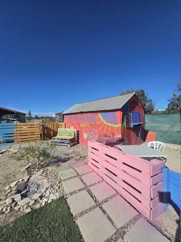 Un pequeño cobertizo de colores con la etiqueta "Farm House" se alza en un patio con vallas de palés pintados, una mesa con sillas, adoquines, bajo un cielo azul despejado.