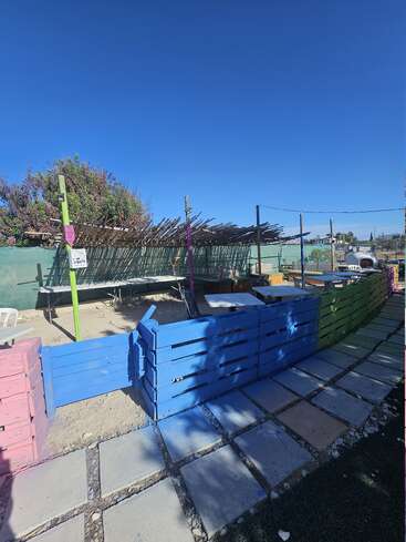 Esta imagen muestra una zona al aire libre con vallas de madera de colores, mesas y asientos. Hay una sombra de bambú en lo alto y escalones adoquinados bajo un cielo azul despejado.