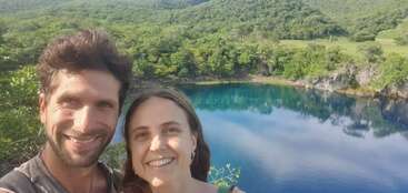 Una pareja sonriente se toma un selfie al aire libre con un hermoso lago azul y exuberantes colinas verdes boscosas de fondo, disfrutando de un día soleado y tranquilo en la naturaleza.