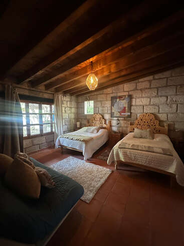 Esse quarto aconchegante tem duas camas de solteiro de madeira ornamentada, uma janela grande, sofá, iluminação aconchegante, paredes de pedra rústica, vigas de madeira no teto e um tapete branco macio.