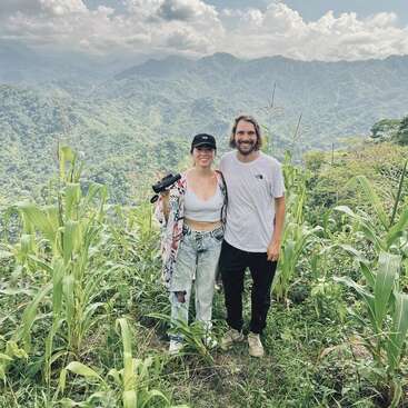 A smiling couple stands together in a lush, green field with tall plants, enjoying a scenic mountain landscape under a cloudy sky on a bright day.