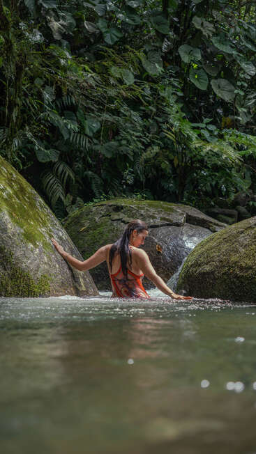 A woman in a red swimsuit wades through a clear, shallow stream surrounded by mossy rocks and lush, dense tropical jungle vegetation. Peaceful, serene moment.
