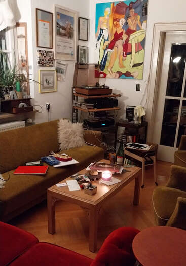 A cozy living room with a mustard couch, art on the walls, a record player, books, a candle, wine bottle, and scattered papers on the table.