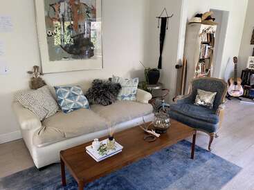 L'image représente un salon confortable avec un canapé blanc, un fauteuil bleu, une table basse en bois et un tapis bleu, avec divers objets décoratifs et une guitare en arrière-plan.
