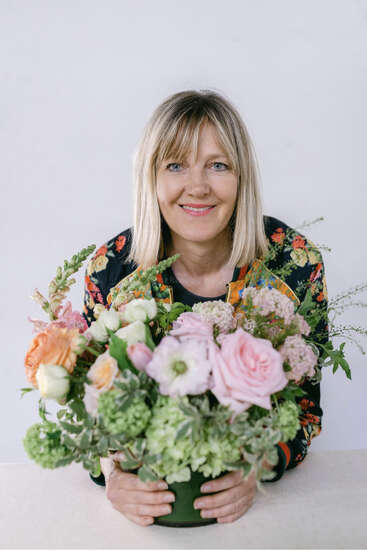 L'image représente une femme aux cheveux blonds, tenant une composition florale vibrante composée de roses roses et d'autres fleurs, sur un fond blanc.