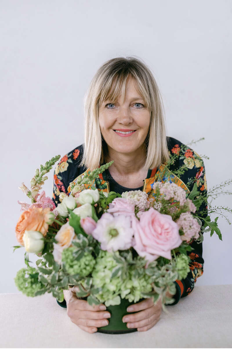 L'image représente une femme aux cheveux blonds, tenant une composition florale vibrante composée de roses roses et d'autres fleurs, sur un fond blanc.