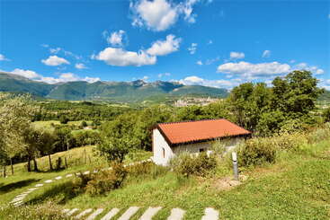 Une petite maison blanche au toit rouge est nichée sur une colline verdoyante, entourée d'arbres, surplombant des montagnes lointaines et un ciel bleu clair et lumineux.