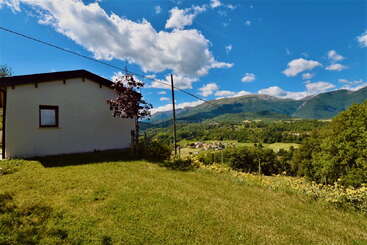 Une petite maison se dresse sur une colline verdoyante surplombant une vallée luxuriante, parsemée d'arbres, sous un ciel bleu éclatant parsemé de nuages blancs cotonneux.