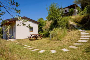 Une petite maison blanche avec une table de pique-nique en bois se trouve sur une pelouse verdoyante. Un chemin en pierre serpente jusqu'à une autre maison entourée d'arbres et de verdure.