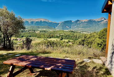 Une table de pique-nique en bois est posée sur un terrain herbeux, surplombant une vallée pittoresque remplie de champs verts, d'arbres et de montagnes lointaines sous un ciel bleu clair.