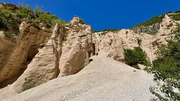 De imposantes formations rocheuses couleur sable s'élèvent abruptement sous un ciel d'un bleu éclatant. Une végétation clairsemée s'accroche aux falaises, contrastant avec le paysage aride et rocheux en contrebas.