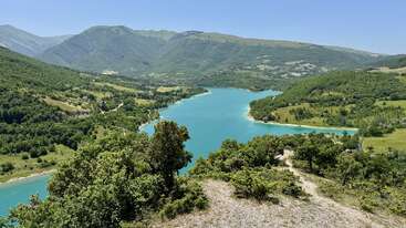 Une rivière sinueuse aux eaux turquoise traverse une vallée verdoyante entourée de collines ondulantes et de montagnes lointaines, sous un ciel bleu limpide par une journée ensoleillée.