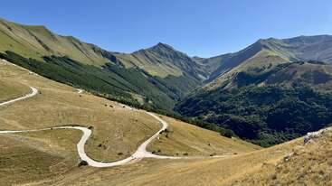 Une route sinueuse traverse des collines verdoyantes et dorées, menant vers des montagnes luxuriantes sous un ciel bleu limpide. La lumière du soleil baigne magnifiquement ce paysage paisible et vaste.