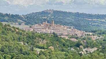 Cette image montre une ville pittoresque perchée au sommet d'une colline, entourée de collines verdoyantes et de maisons éparpillées. Des bâtiments historiques en pierre dotés de tours dominent ce paysage charmant et paisible.