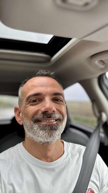 A man with short gray hair and a neatly trimmed beard is smiling while sitting in a car, wearing a white shirt and a seatbelt.