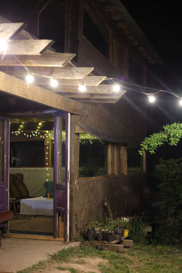 Une maison rustique de nuit se caractérise par des guirlandes lumineuses à l'extérieur, des portes ouvertes, des plantes en pot visibles et un intérieur douillet avec d'autres lumières, des chaises et une table couverte.