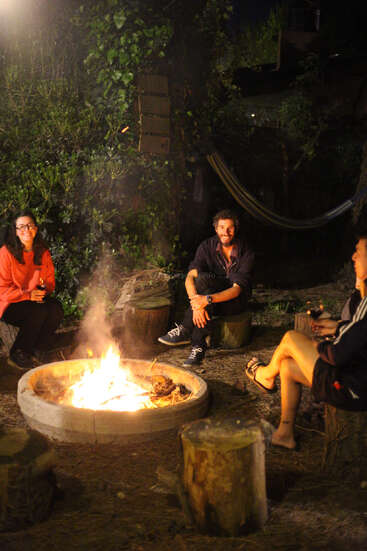 Trois amis sont assis autour d'un feu de camp la nuit, discutant et buvant du vin. Entourés par la nature, un hamac est suspendu à l'arrière-plan, créant une atmosphère confortable.