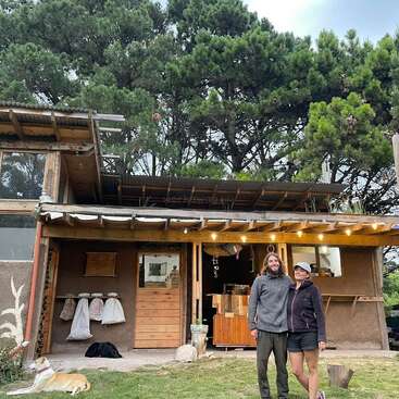 A imagem mostra uma cena serena de um casal em frente a uma aconchegante casa de madeira, cercada por árvores exuberantes e um cachorro deitado na grama.