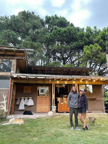 L'image représente une scène sereine d'un couple se tenant devant une maison en bois confortable, entourée d'arbres luxuriants et d'un chien couché dans l'herbe.