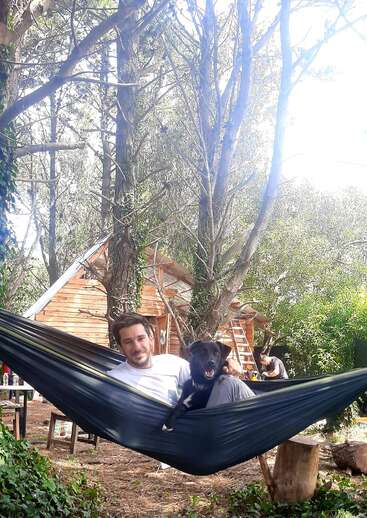 L'image représente un homme assis dans un hamac avec un chien, sur fond de maison en bois et d'arbres.