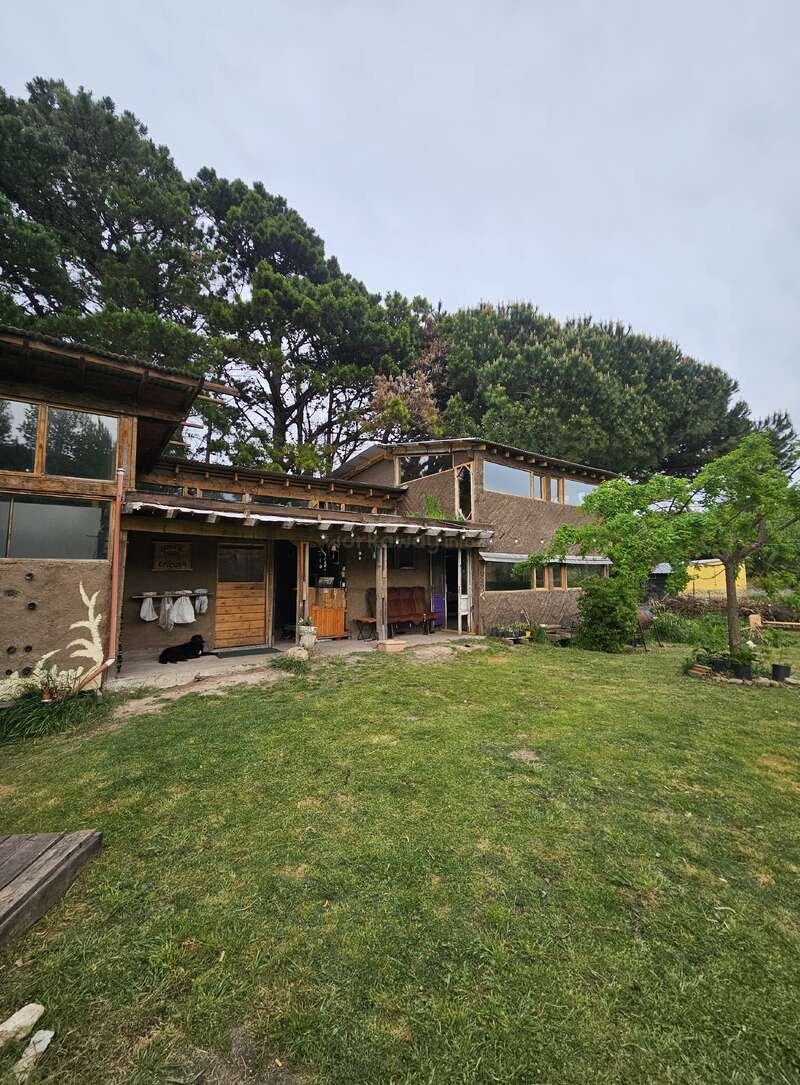 Cette image montre une maison rustique avec des matériaux naturels, entourée de verdure et d'arbres. Il y a un porche, une pelouse, des éléments en bois et une atmosphère paisible et rurale.