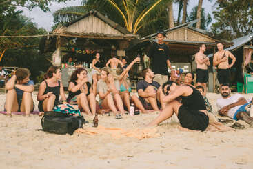 A group of friends relax on a sandy beach, laughing and talking. Behind them are rustic beach huts, palm trees, and more people enjoying the tropical atmosphere.