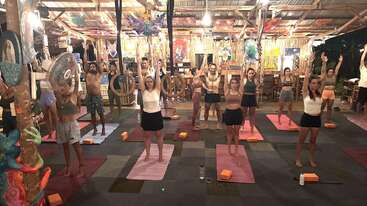 A group of people practice yoga indoors on colorful mats, standing with arms raised. The setting is vibrant with bright lights, artistic decor, and a relaxed atmosphere.