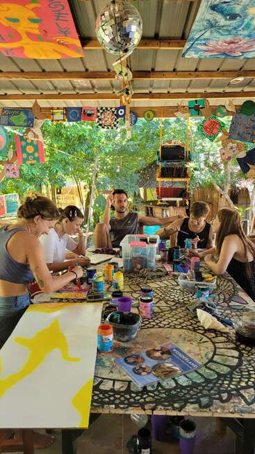 Five people sit around a colorful, art-covered table, painting and chatting in a lively outdoor studio decorated with hanging artwork, greenery, and a disco ball.