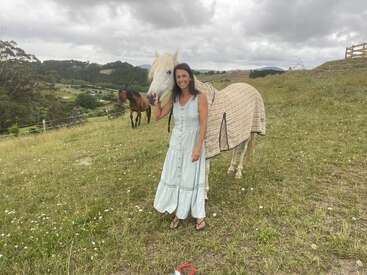A woman in a light blue dress stands on a grassy hill beside a white horse wearing a blanket, with another horse grazing nearby under cloudy skies.
