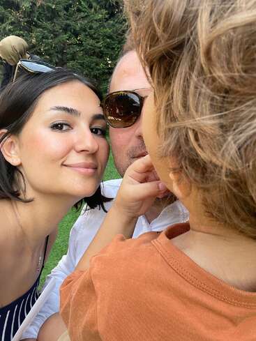 A woman, a man wearing sunglasses, and a child close together outdoors, smiling. Lush greenery in the background, suggesting a happy moment captured in nature.