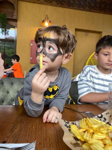 A young boy with a Batman face paint holds a crinkle-cut fry, looking sideways, while sitting at a table with another child and a plate of fries.