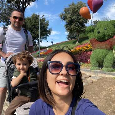 Eine glückliche Familie genießt einen sonnigen Tag im Freien. Eine Frau lächelt im Vordergrund, während ein Mann und ein Kind im Kinderwagen sitzen. In der Nähe wächst ein grüner Bär.