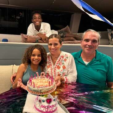 L'image montre une famille de quatre personnes célébrant un anniversaire, avec un gâteau et des bougies sur une table, et un homme et une femme assis sur un canapé derrière eux.