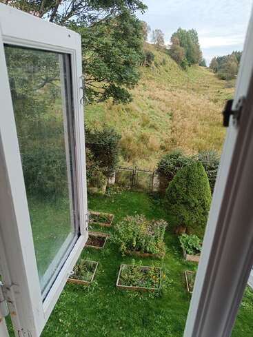 Das Foto zeigt ein offenes Fenster mit Blick auf einen üppigen Garten mit Hochbeeten, grünem Rasen, Bäumen, einem Hügel und fernen Feldern unter einem bewölkten Himmel.