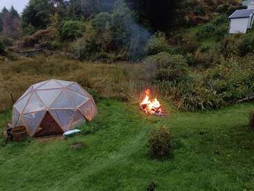 Ein geodätisches Kuppelgewächshaus und ein brennendes Lagerfeuer stehen auf saftig grünem Gras, umgeben von dichter Vegetation und Bäumen, mit einem weißen Haus in Sichtweite.