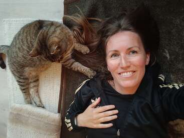 Eine Frau liegt auf einem dunklen Teppich und lächelt in die Kamera. Neben ihr berührt eine getigerte Katze sanft ihr langes Haar mit ihrer Pfote. Gemütlicher Moment.