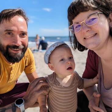 The image shows a family of three posing for a selfie on a beach, with the father, mother, and baby dressed in casual attire and smiling at the camera.