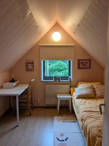 This cozy attic bedroom features a sloped ceiling, a single bed with yellow bedding, a white desk and chair, window with lush green view, minimalist décor.